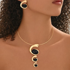 Pode incluir: Conjunto de joias douradas com colar e brinco a condizer. O colar tem um pendente curvo com três pedras ovais pretas. O brinco é um design curvo com uma pedra oval preta.