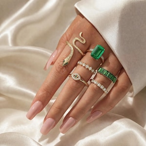 May include: A selection of gold rings on a hand. The rings feature a snake design, an emerald-cut green gemstone, and rings with clear and green gemstones. The hand displays long, polished nails.