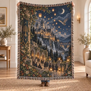 May include: A woven throw blanket featuring a detailed fantasy scene of a castle on a cliff at night, illuminated by glowing lanterns and a crescent moon. The design includes a stone archway, a tree banner, and an owl flying in the starry sky.