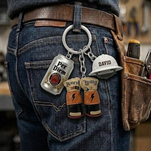 Op de afbeelding: Een metalen sleutelhanger aan een broeklus met een zilverkleurige bierblikje bedel met de tekst Per Diem, een witte veiligheidshelm met de naam David, en twee beige en zwarte werkhandschoen bedels met de tekst Honest Living erop.