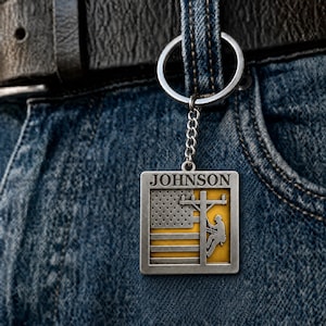 Puede incluir: Un llavero cuadrado de metal con el nombre JOHNSON en la parte superior. El diseño incluye un motivo de bandera estadounidense en tono plateado combinado con un fondo amarillo y la silueta de un trabajador subiendo un poste eléctrico, con anilla.