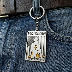 Op de afbeelding: Een rechthoekige zilverkleurige metalen sleutelhanger met de naam JOHNSON bovenaan, voorzien van een silhouet van een brandweerman gecentreerd over een gestileerd Amerikaans vlagontwerp. Het ontwerp bevat een gele bliksemschicht.