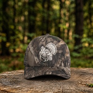 Könnte beinhalten: Eine Camouflage-Baseballkappe mit einer weißen Stickerei eines Truthahns auf der Vorderseite. Die Kappe hat eine strukturierte Krone und einen gebogenen Schirm und zeigt ein waldinspiriertes Muster in Braun-, Grün- und Schwarztönen.