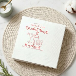 May include: A square white paper napkin featuring red printed text that reads Welcome to ISABELLA'S Bridal Bash Weekend CAPE COD, MA | SEPTEMBER 2025, accompanied by a line art illustration of a martini glass, a liquor bottle, and two glasses.