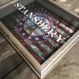 May include: A wooden shadow box with a glass front. The artwork inside features an American flag design with the words "STAN SPERRY" in white, and an emblem that reads "ARMY NATIONAL GUARD". The frame has a rustic, weathered appearance.