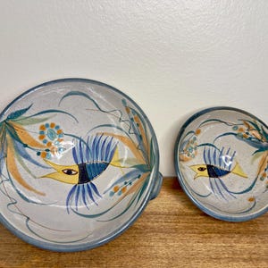 May include: Two round ceramic bowls with a speckled off-white glaze, featuring hand-painted designs of yellow and blue fish surrounded by teal and orange foliage. The larger bowl includes a small side handle, and both have blue painted rims.
