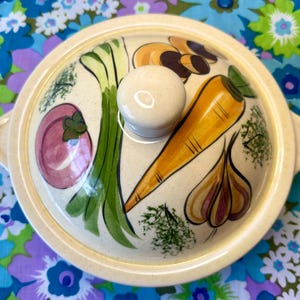 May include: Cream-colored ceramic casserole dish with a lid featuring hand-painted vegetables. The lid has a white knob and illustrations of a carrot, onion, garlic, and other vegetables. The dish has two side handles.