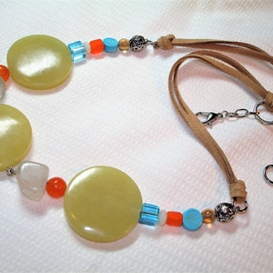 May include: A necklace featuring large, round, pale yellow beads interspersed with smaller, colorful beads in orange, blue, and clear. The necklace has a tan suede cord with a silver-tone clasp and rings.