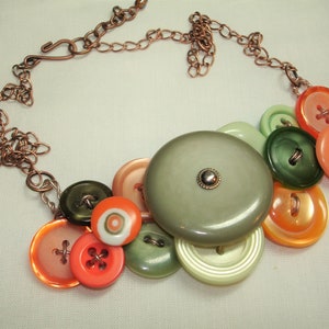 May include: A handmade necklace featuring a cluster of colorful buttons in shades of orange, green, and pale green. The buttons are various sizes and shapes, with a large, round, gray button at the center. The necklace has a copper chain.