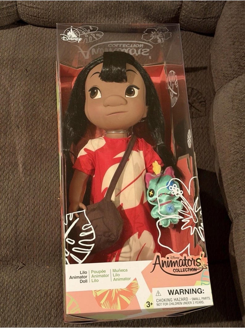 Puede incluir: Mu&ntilde;eca Lilo de la colecci&oacute;n Disney Animators en una caja transparente. La mu&ntilde;eca tiene cabello negro, ojos marrones y viste un vestido rojo con estampados florales blancos. Un bolso marr&oacute;n cuelga de su hombro y se incluye un peluche de Stitch.