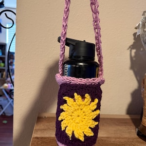 May include: A purple crochet water bottle holder with a yellow sun design. The holder has a pink trim and a matching pink and purple strap. A black water bottle is inside the holder.