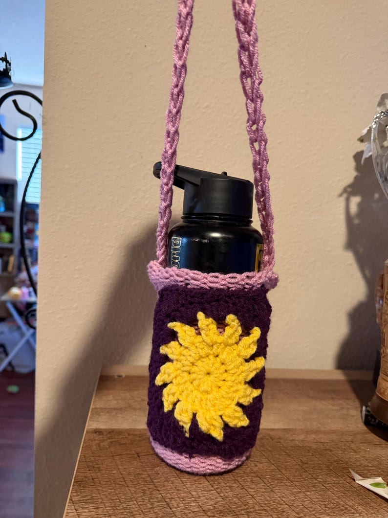 May include: A purple crochet water bottle holder with a yellow sun design. The holder has a pink trim and a matching pink and purple strap. A black water bottle is inside the holder.