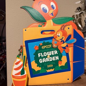May include: A rectangular orange and yellow bag featuring an Orange Bird character on top and a bee character on the front. The center displays a blue-bordered sign reading EPCOT INTERNATIONAL FLOWER GARDEN FESTIVAL 2026. A small ice cream charm hangs from the side.