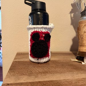 May include: A black water bottle with the word "World" printed on it, in a white and red crocheted cosy. The cosy features a black Mickey Mouse design. The bottle is on a wooden surface.