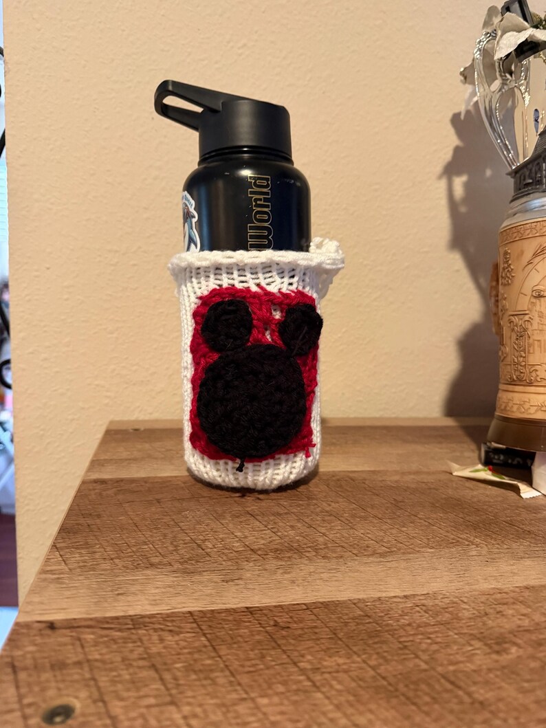 May include: A black water bottle with the word "World" printed on it, in a white and red crocheted cosy. The cosy features a black Mickey Mouse design. The bottle is on a wooden surface.