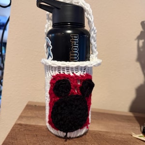 May include: A black water bottle in a white crocheted holder. The holder features a red and black cartoon character design. The bottle has the word "World" printed on it.