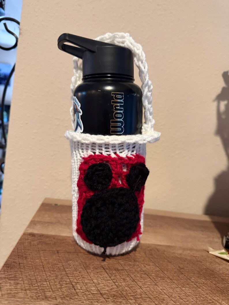 May include: A black water bottle in a white crocheted holder. The holder features a red and black cartoon character design. The bottle has the word "World" printed on it.