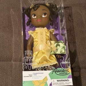 Puede incluir: Mu&ntilde;eca Tiana de la colecci&oacute;n Disney Animator en una caja de pl&aacute;stico transparente. La mu&ntilde;eca tiene piel oscura, cabello negro y viste un vestido amarillo con un lazo a juego. La caja presenta el logotipo de Disney y la inscripci&oacute;n "Animator's Collection".