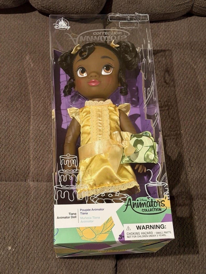 Puede incluir: Mu&ntilde;eca Tiana de la colecci&oacute;n Disney Animator en una caja de pl&aacute;stico transparente. La mu&ntilde;eca tiene piel oscura, cabello negro y viste un vestido amarillo con un lazo a juego. La caja presenta el logotipo de Disney y la inscripci&oacute;n "Animator's Collection".