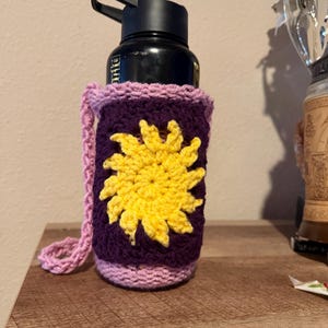 May include: A black water bottle in a handmade crochet cosy. The cosy is purple with a yellow sun design and a pink border and strap. The bottle is on a wooden surface.