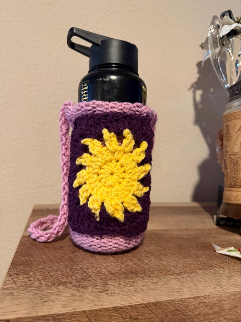 May include: A black water bottle in a handmade crochet cosy. The cosy is purple with a yellow sun design and a pink border and strap. The bottle is on a wooden surface.