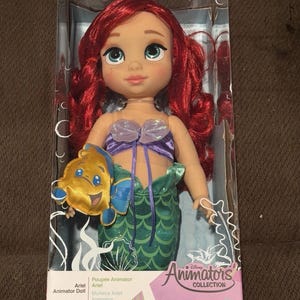 Puede incluir: Mu&ntilde;eca Ariel de la colecci&oacute;n Disney Animators. La mu&ntilde;eca tiene cabello rojo, ojos azules y viste un top morado y una cola de sirena verde. Incluye un peluche de Flounder. Empaquetada en una caja transparente.