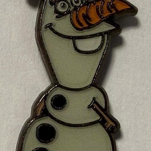 May include: A metal enamel pin featuring the character Olaf, a snowman with a pale off-white body, a bright orange carrot nose, brown twig-like hair and arms, and three black circular buttons on the torso, outlined in a dark metallic border.