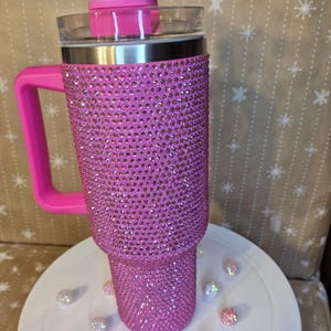 May include: A pink, bedazzled tumbler with a handle and a clear lid. The tumbler is covered in small, sparkling rhinestones. The handle and the lid's slider are also pink. Several decorative rhinestone balls are scattered around the base.