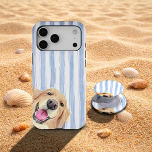 May include: A light blue and white striped phone case featuring a smiling golden retriever illustration. A matching phone grip is also visible. The items are on a sandy surface with seashells.