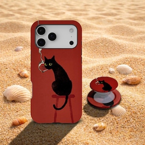 Puede incluir: Una funda de smartphone roja y un soporte circular a juego, ambos con una ilustración gráfica de un gato negro de ojos amarillos sentado en un taburete rojo mientras sostiene un anillo de color plateado en la boca sobre un fondo rojo.
