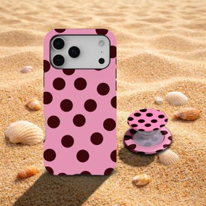 May include: Pink phone case and matching phone grip with a repeating pattern of dark brown polka dots. The phone case is on a sandy surface with seashells.