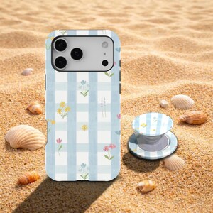 May include: A phone case and phone grip with a light blue and white gingham pattern, adorned with small yellow, pink, and blue floral designs. The items are displayed on sand, with seashells scattered around. The phone case is for a mobile phone.