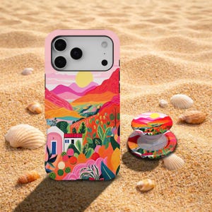 May include: A smartphone case and a matching circular phone grip featuring a vibrant, colourful illustration of a Mediterranean-style landscape with pink mountains, a yellow sun, white buildings, and lush green foliage against a bright orange sky.