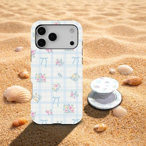 May include: A white phone case and matching phone grip with a floral and bow pattern on a light blue plaid background. The case is on a sandy surface with seashells.