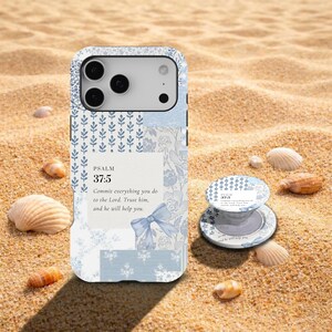 May include: A light blue and white phone case with a floral and bow design, featuring the text "PSALM 37:5". A matching phone grip is also visible. The items are on a sandy surface with seashells.