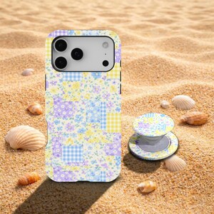 Pode incluir: Uma capa de telefone e um suporte a condizer com um design de patchwork floral, pousados na areia com conchas. A capa e o suporte apresentam uma mistura de padrões florais e xadrez pastel em amarelo, azul e roxo. O suporte é circular.