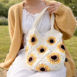 Op de afbeelding: Een witte gehaakte draagtas met een zonnebloemontwerp. De tas heeft gele en bruine zonnebloemen op een witte achtergrond. Het heeft een schouderriem. De tas wordt vastgehouden door een persoon die een gele cardigan en een witte jurk draagt.