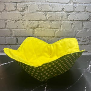 May include: A square bowl with a bright yellow interior and a green and white checkered exterior. The bowl is sitting on a black and white marble surface, with a white brick wall in the background.