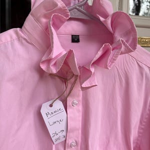 May include: A light pink button-down shirt featuring a prominent ruffled collar, a front pocket, and a white paper tag attached with twine. The tag displays the handwritten text 'Maeve', 'Large', and '23 -> 25'. A black inner label shows '165/102A 44'.