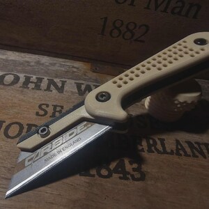 Puede incluir: Un cuchillo utilitario beige y negro con una hoja plateada. El cuchillo tiene un mango texturizado y el texto "CARBIDE MADE IN ENGLAND" es visible en la hoja. El cuchillo está abierto y descansa sobre una superficie de madera con texto grabado.