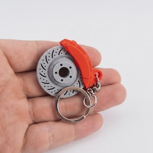 Peut inclure: Un porte-clés avec un disque de frein gris et rouge. Le disque est gris avec des trous percés et un moyeu central. Un étrier de frein rouge est attaché. Le porte-clés comprend un anneau fendu argenté. Accessoire sur le thème automobile.