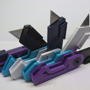 Può includere: Una collezione di taglierini in vari colori, tra cui viola, blu, bianco e nero. Ogni taglierino ha una lama affilata argentata che si estende da un alloggiamento colorato. I taglierini sono disposti in fila, mostrando il loro design.