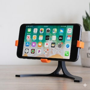 Puede incluir: Un teléfono inteligente negro se muestra en un soporte para teléfono negro y naranja. La pantalla del teléfono muestra iconos de aplicaciones. El soporte tiene una base curva y soportes naranjas. La hora es 12:26 PM.