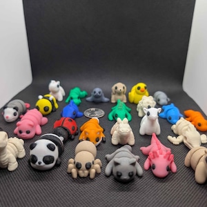 May include: A collection of small, colorful clay animal figurines. The assortment includes a panda, ladybug, axolotl, and other creatures in various colors. The figurines are arranged on a dark surface.