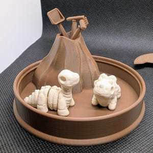 Puede incluir: Un diorama de dinosaurios marrón impreso en 3D con un volcán con herramientas, un dinosaurio de cuello largo y un triceratops. Los dinosaurios son de color blanquecino. El diorama está en una base circular marrón.
