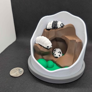 May include: A white terrarium-style container with a grey base, featuring a miniature brown landscape with green accents and three small, stylized insect figures. A UK coin is next to the terrarium.