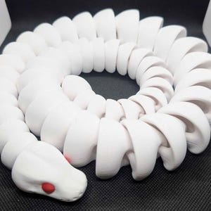 May include: A white, articulated snake toy with a rounded head and red eye details. The snake is coiled, showcasing its segmented body. The toy is made of a smooth, solid material.