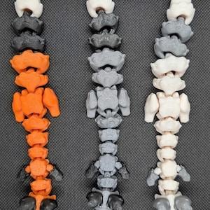 Könnte beinhalten: Drei bewegliche Wirbelsäulenmodelle in Orange, Grau und Weiß, jeweils mit schwarzen und grauen Akzenten. Die Modelle sind vertikal vor einem dunkelgrauen Hintergrund angeordnet und zeigen ihr segmentiertes Design und anatomische Details.