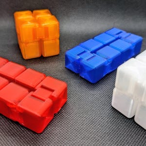 Könnte beinhalten: Vier farbenfrohe Zappelspielzeuge in Orange, Blau, Rot und Weiß. Jedes Spielzeug ist ein Würfel aus kleineren, verbundenen Blöcken. Die Spielzeuge sind auf einer dunkelgrauen Oberfläche mit weißem Hintergrund angeordnet.