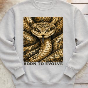 May include: A light grey crewneck sweatshirt featuring a large, centred graphic of a coiled rattlesnake with brown and tan scales, a visible tongue, and the text BORN TO EVOLVE printed in dark grey capital letters directly below the snake image.